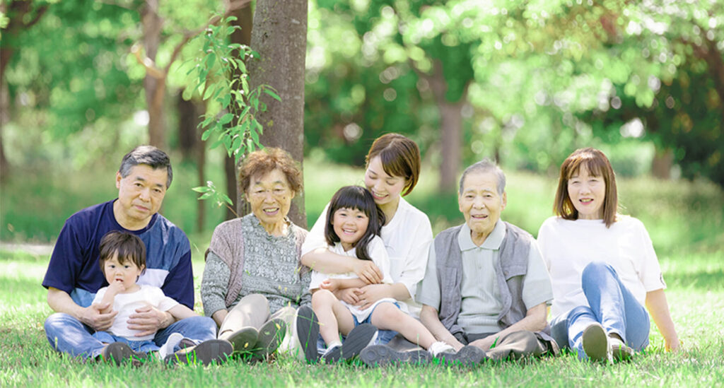 芝生の上でくつろぐ三世代の家族。笑顔があふれ、温かい家族のつながりが感じられる。