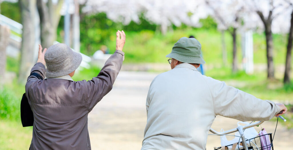 公園の遊歩道で帽子をかぶったシニア夫婦が、自転車を押しながら楽しそうに会話している様子