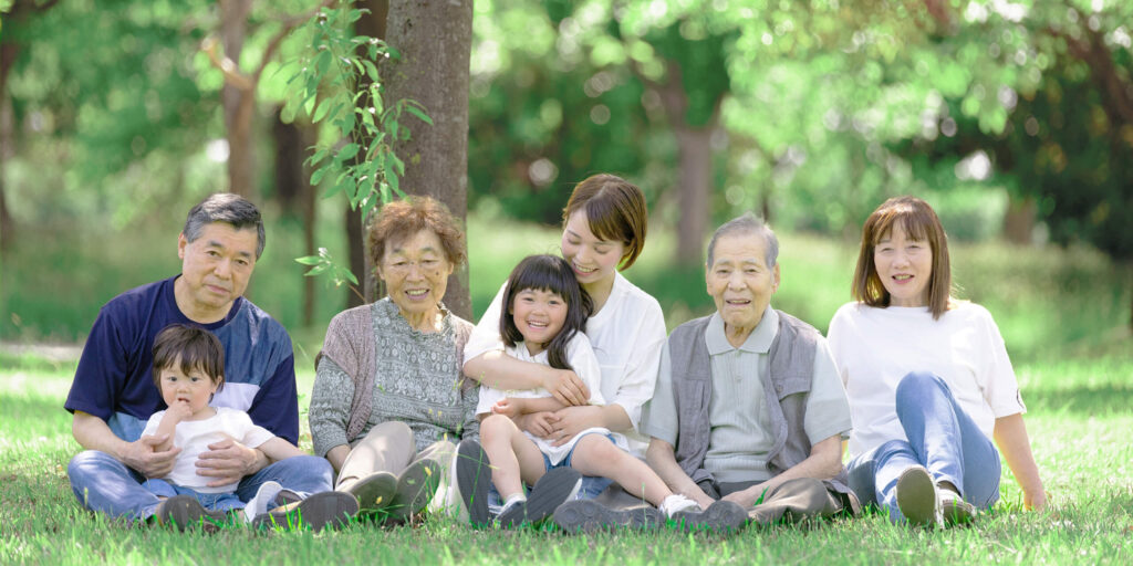 芝生の上でくつろぐ三世代の家族。笑顔があふれ、温かい家族のつながりが感じられる。